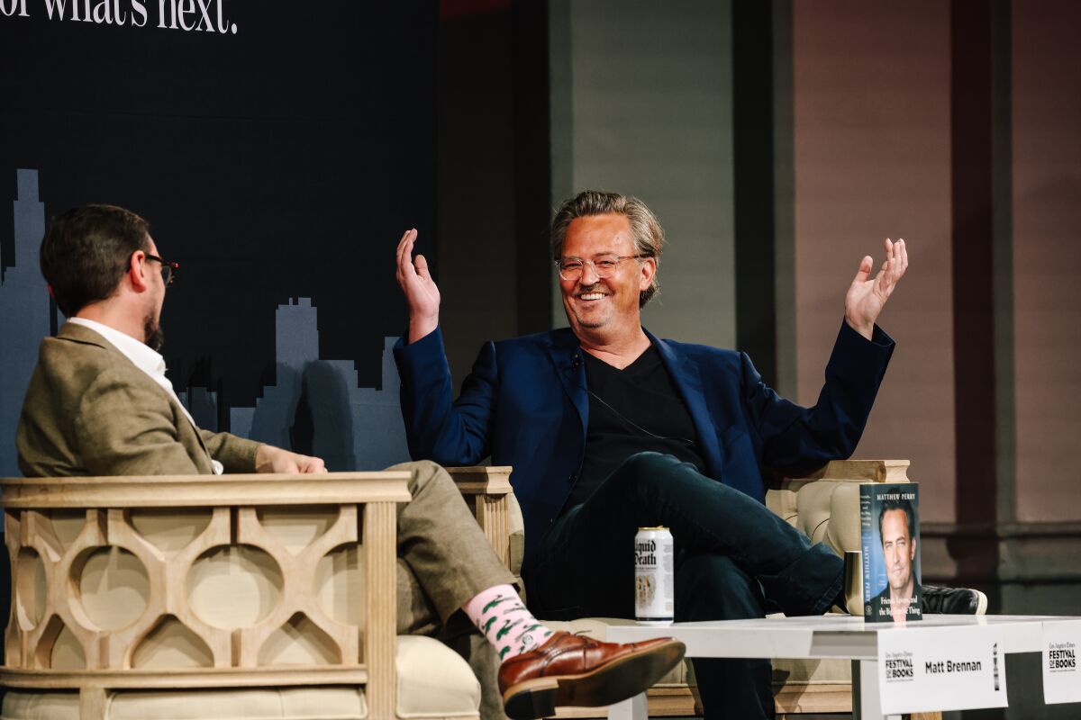 A smiling Matthew Perry raises his arms into the air while seated onstage, talking with an interviewer