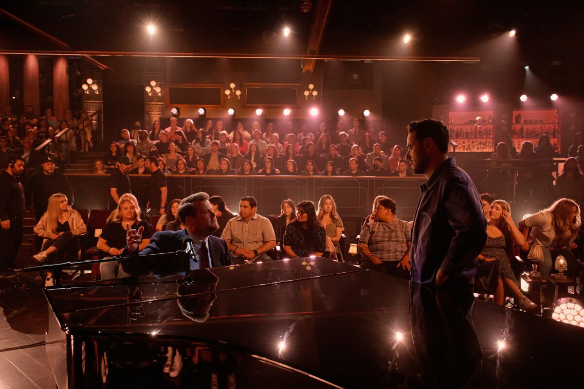 James Corden singing at a piano with a studio audience behind him.