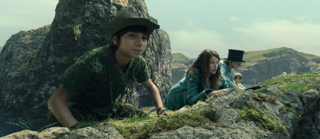 Peter Pan (Alexander Molony), Wendy (Ever Anderson), John Darling (Joshua Pickering), and Michael Darling (Jacobi Jupe) crouch at the top of a mossy, rocky ridge, looking over the edge in Peter Pan &amp; Wendy