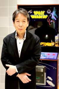 Tomohiro Nishikado posing next to a Space Invaders arcade cabinet