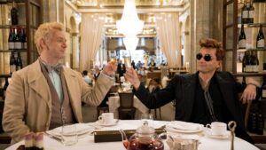 in the middle of a crowded restaurant, Aziraphale, dressed in lighter colors, and Crowley, dressed in mostly black, have a toast on Good Omens