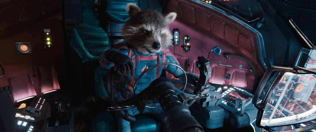 Anthropomorphic raccoon Rocket (body acted by Sean Penn, voiced by Bradley Cooper) sits in the cockpit of a spaceship in full Guardians of the Galaxy blue-and-red jumpsuit uniform in Guardians of the Galaxy Vol. 3