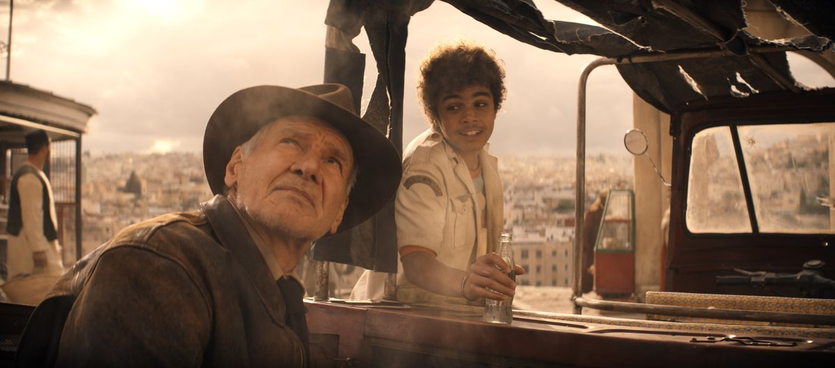 Indiana Jones (Harrison Ford) looks up at an overcast grey sky while a grinning teenager (Ethann Isidore) tries to talk to him as he gears up for a ride in his truck in Indiana Jones and the Dial of Destiny