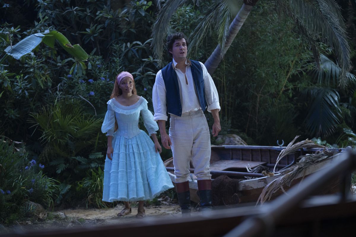 Eric and Ariel in the live-action Little Mermaid stand on a beach. Ariel wears a light blue dress, while Eric wears a white shirt with a blue vest and khaki trousers.