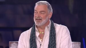 Ray Stevenson in a white shirt and scarf with a beard sitting
