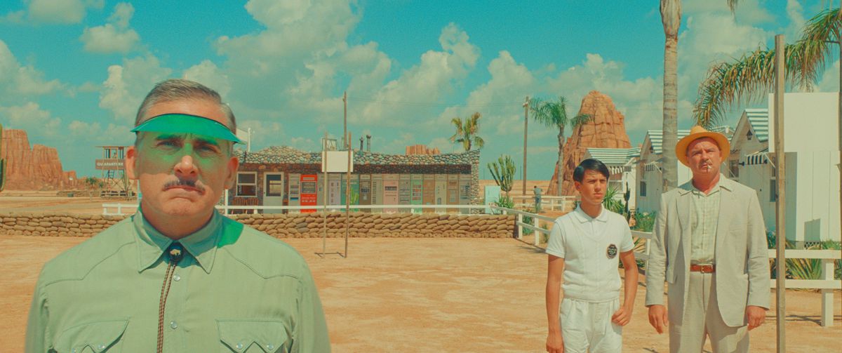 A motel manager (Steve Carell) stands in front of a desert motel, wearing a green sunshade and looking into the camera, while two people in pale clothing (Aristou Meehan and Liev Schreiber) stand in the distance behind him in Wes Anderson’s Asteroid City