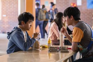 Ben Wang and Jim Liu as Jin Wang and Wei-Chen sit across from each other at a school lunch table. Jin has a white bread sandwich and apple juice, while Wei-Chen has metal tin of food and is eating with chopsticks.