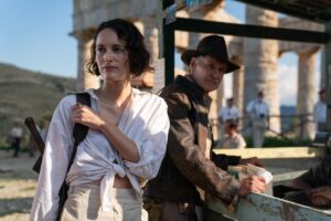 Helena Shaw (Phoebe Waller-Bridge, in a white button-up shirt tied up to the navel) stands in front of some ruins with Indiana Jones (Harrison Ford, wearing his classic Indiana Jones outfit) in Indiana Jones and the Dial of Destiny.