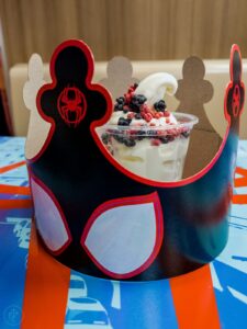 Burger King’s Spider-Verse Sundae, a swirl of soft-serve vanilla ice cream topped with red and black popping candies in a cup, sitting inside a Spider-Verse-branded cardboard crown on a table with Spider-Man artwork