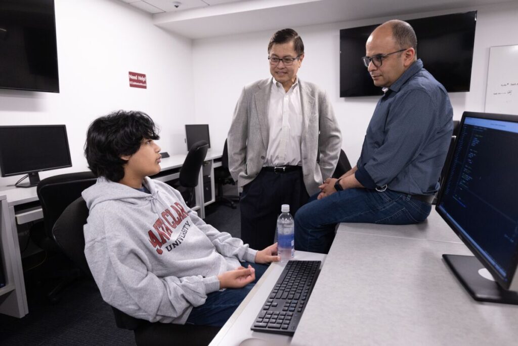 Kairan Quazi, seated behind a desk with a computer screen, talks with two men.