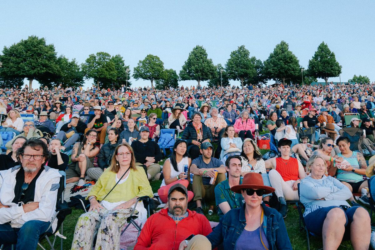 A large audience sits on chairs and the grass outdoors.