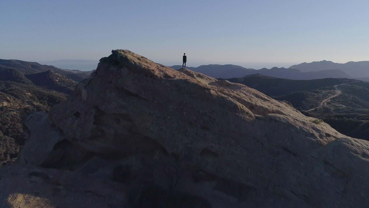 Narcissa Wright as a tiny figure in silhouette, standing on a vast tilted rock overlooking some low mountains in a shot from Break the Game