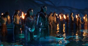 A group of Na’vi gather at night for a ceremony, standing knee-deep in water and holding torches, with Na’vi played by Kate Winslet and Cliff Curtis presiding, in Avatar: The Way of Water