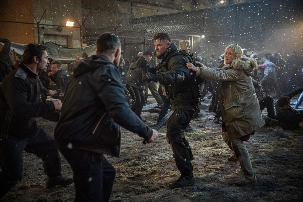 Chris Hemsworth holds his fists up as a blonde woman (Tinatin Dalakishvili) hides behind him in a snowy fight scene in Extraction 2.