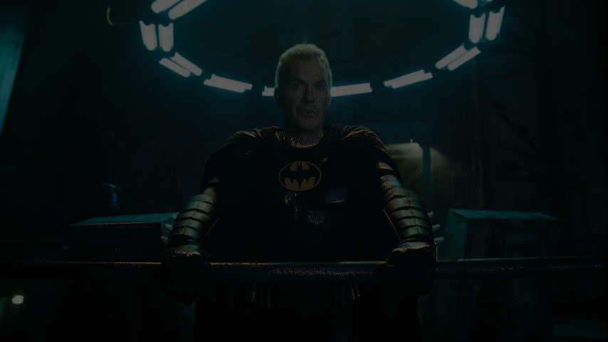 A gif with a slow zoom in on Michael Keaton in the Batman suit (but no cowl), standing at a railing in the Batcave with a halo of fluorescent lights above him in The Flash