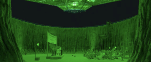 A spaceship largely made of glass hovers above a crowd of people seated in an asteroid blast crater in a shot lit only by eerie green light in Wes Anderson’s Asteroid City