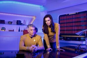 Anson Mount as Captain Pike as Rebecca Romijn as First Officer Una “Number One” Chin-Riley sit and stand behind a desk on the Enterprise.