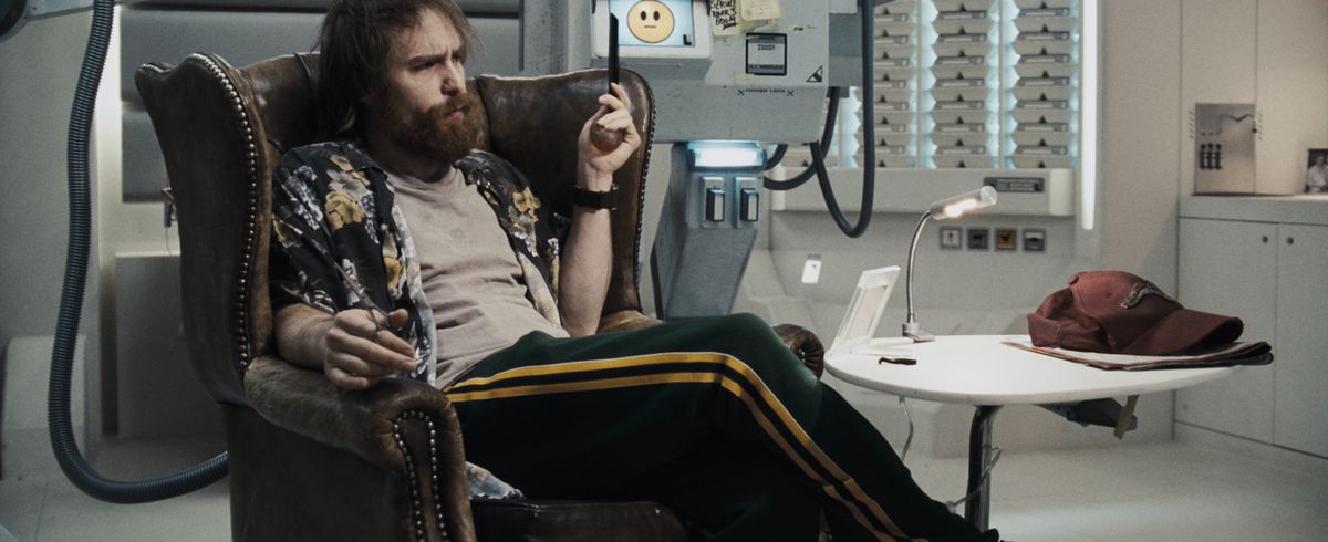 Astronaut Sam Bell (Sam Rockwell), with a big bushy beard, unkempt hair, and stains on his grubby T-shirt, slouches in an antique-looking leather chair amid sleek white space-station surfaces and a boxy robot sporting a no-expression smiley emoji in Duncan Jones’ Moon