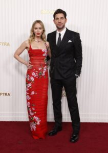 Emily Blunt wears a long red dress and John Krasinski poses in a suit on a red carpet