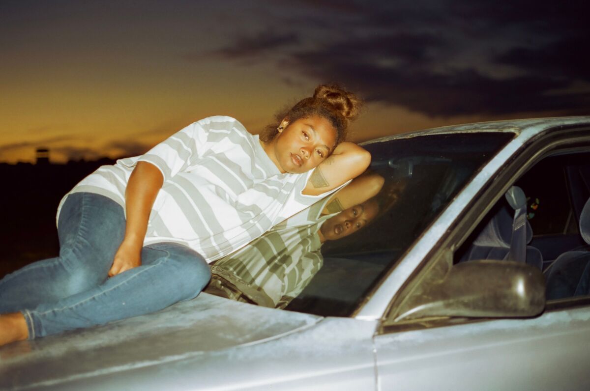 A woman lies on the hood of a car, leaning against the windshield