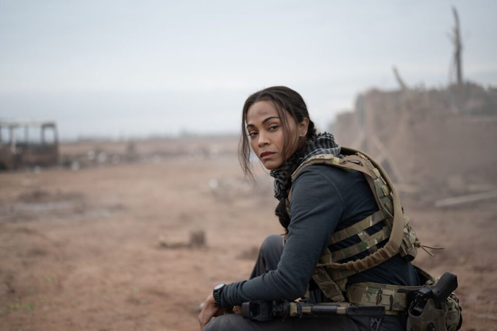 A woman in dark clothing and a camo vest sits on the ground in the desert.