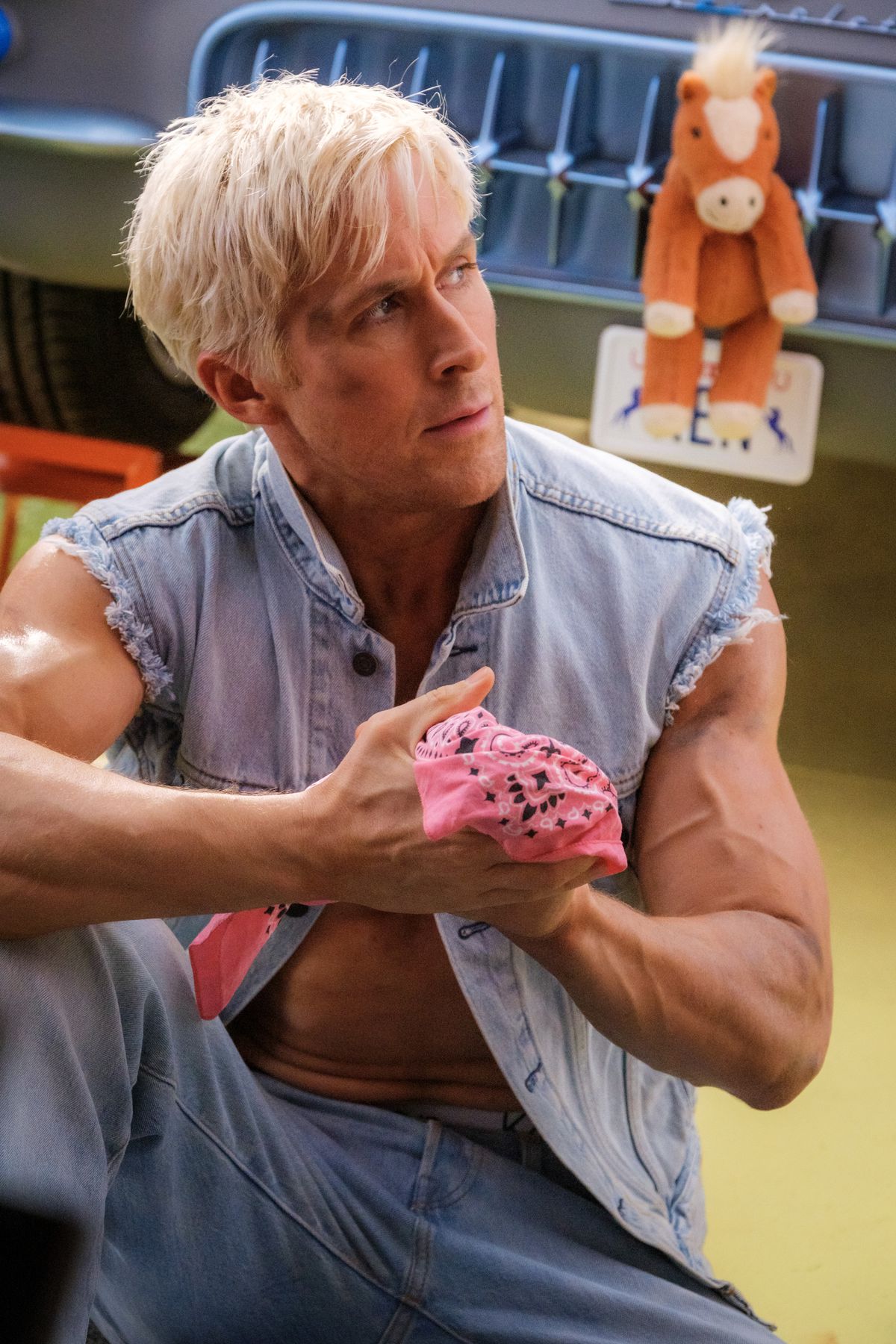 Ryan Gosling as Ken in a denim vest and jeans, a little pink bandana in his hands. He sits on the floor. Behind him there is a truck, with a stuffed horse attached to the front. 