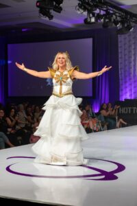 Ashley Eckstein walks down the runway in a white dress with her arms spread open.