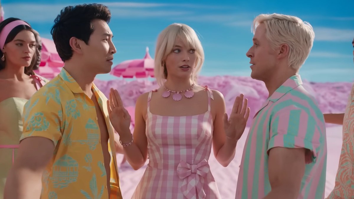 Margot Robbie's Barbie in a pink and white dress stands between Simu Liu and Ryan Gosling's Kens on the beach