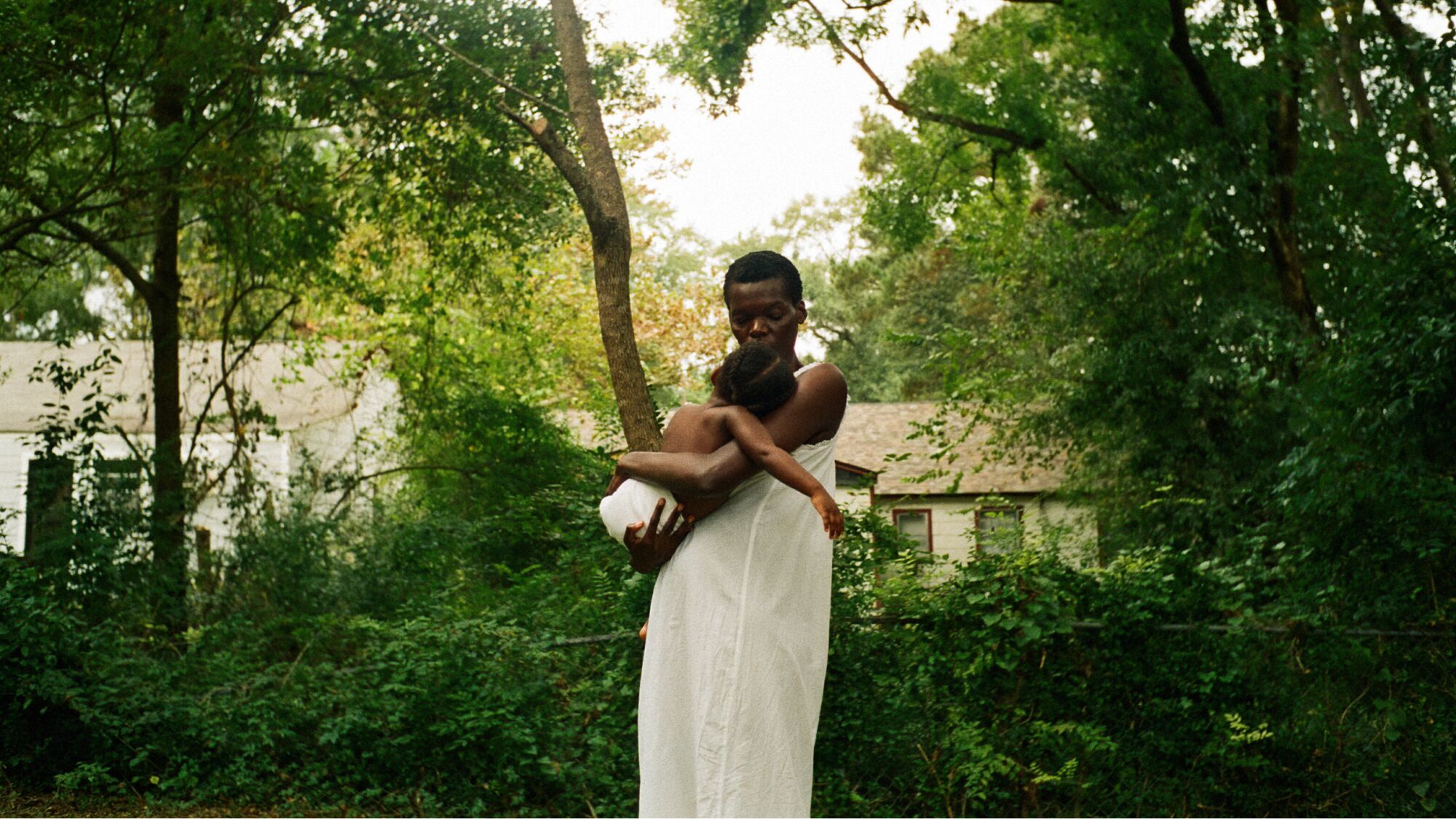 A woman in a white dress in a grove of trees holds a child.