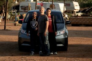 Three men stand in front of a minivan in a dirt parking lot.