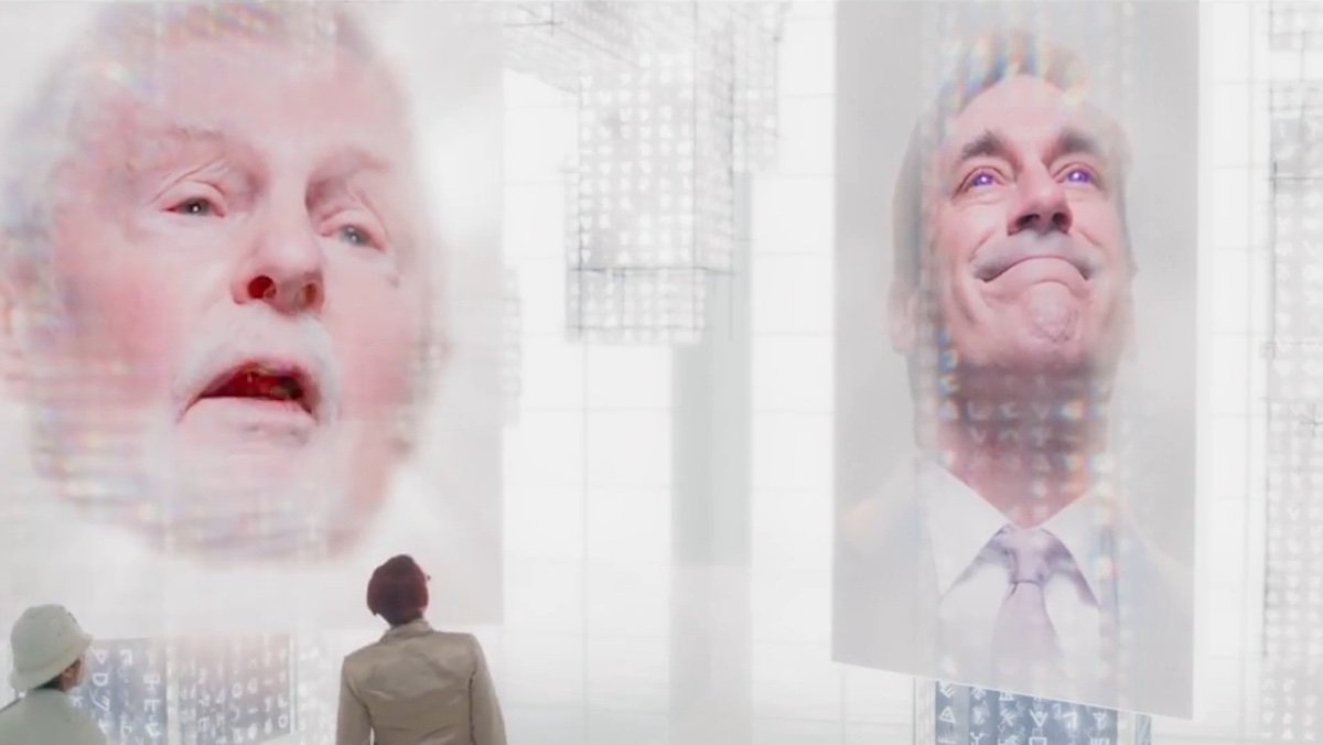 Two people look p at giant screens with heads on Good Omens