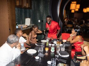 Kevin Hart in a bright red outfit standing and smiling at people sitting at a dinner table