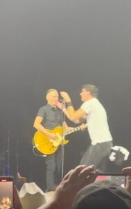 Bryan Adams with guitar on stage man in white shirt grabbing microphone.