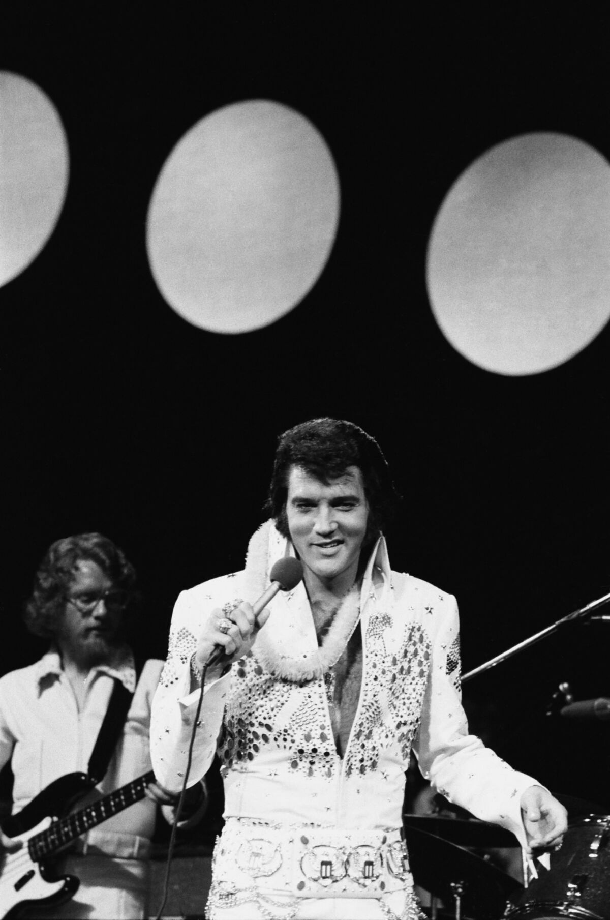 Elvis Presley in a white jumpsuit holding a microphone to his mouth.