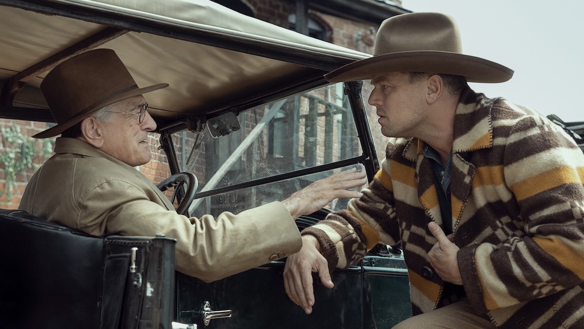 Robert De Niro in a hat and eyeglasses sits in a car speaking to Leonardo DiCaprio also in a hat leaning on the car in Killers of the Flower Moon