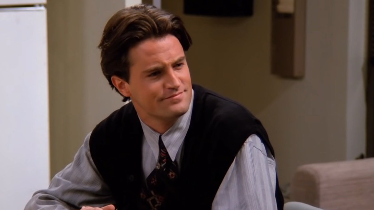 Matthew Perry as Chandler Bing in a black vest and tie sitting on Friends