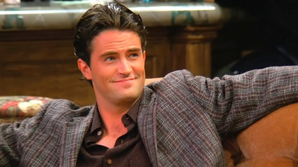 Matthew PErry's Chandler Bing smirks sitting on a couch on Friends