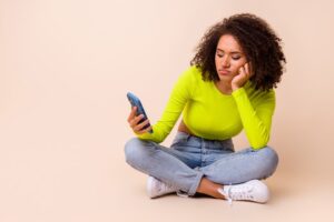 Woman in yellow top and blue jeans, white shoes sitting cross-legged with phone and disapproving expression.
