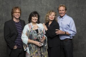 Bill Mumy, Angela Cartwright, Marta Kristen and Mark Goddard stand next to each other in front of a gray backdrop.