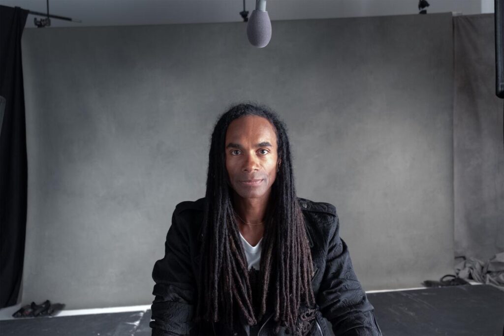 Fab Morvan sits in front of gray backdrop in a white shirt and black jacket with his dreadlocks over his chest.