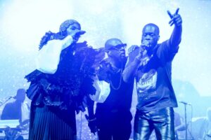 Lauryn Hill, Wyclef Jean and Pras perform onstage with mics held to their mouths as the three embrace