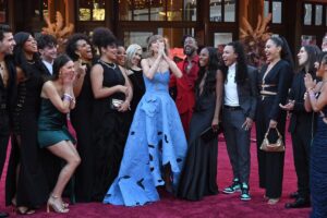 A woman in a blue gown is surrounded by colleagues on a red carpet.