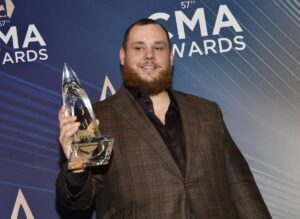 Luke Combs in a suit smiling and looking to his side while holding a pointy, clear award