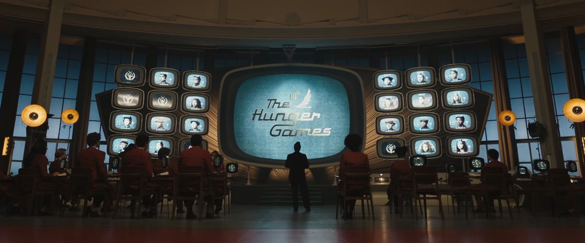 The in-world logo for the Hunger Games on retro TV screens while employees of the games mull about