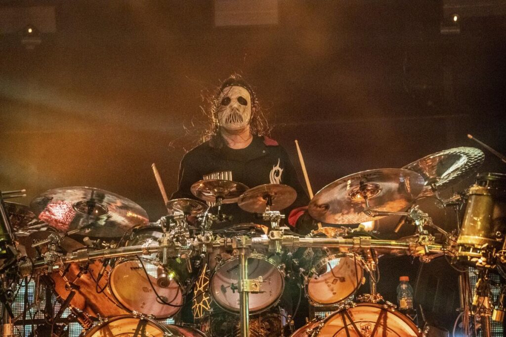 A man with long hair, black and white face paint and a black shirt plays at a drum kit onstage