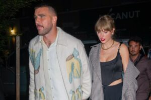 Travis Kelce is wearing a jacket and white shirt and is holding hands with Taylor Swift who is in a plaid coat