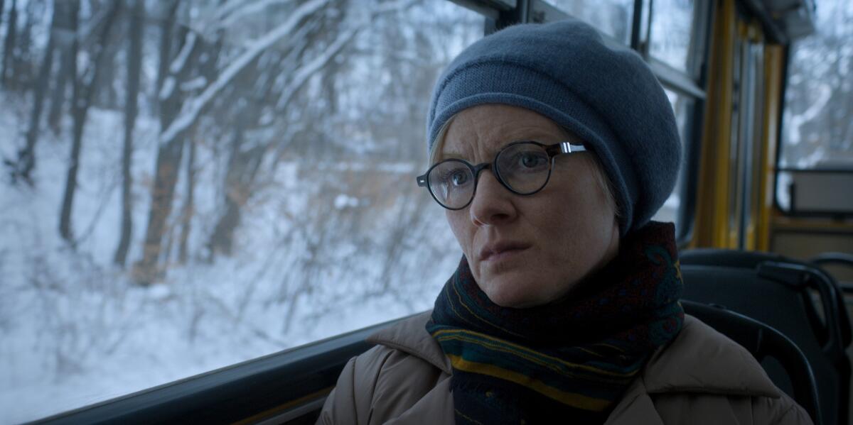 A woman in a gray hat, scarf and glasses sits in car that's moving past a snowy forest.