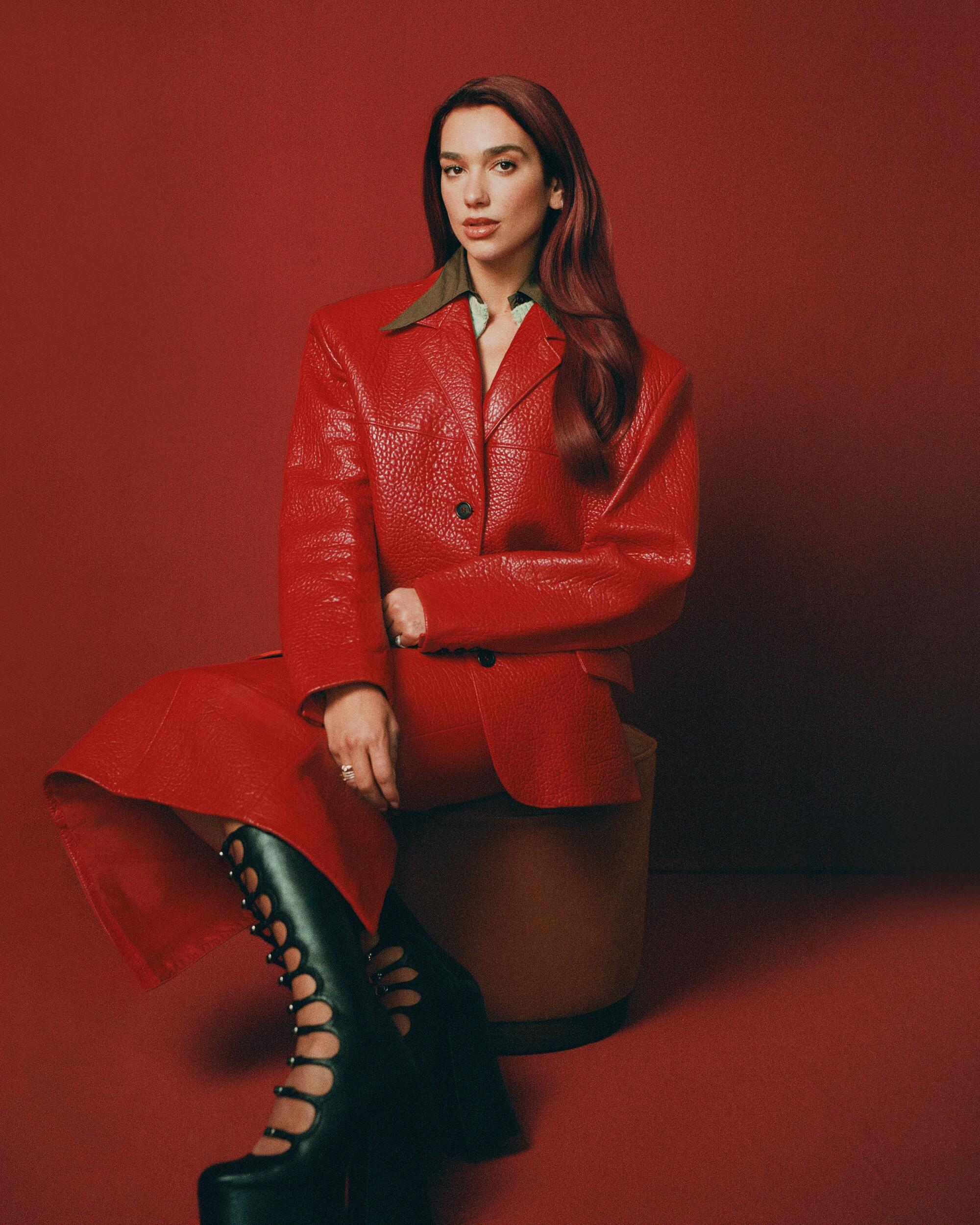Dua Lipa wears a red outfit against a red backdrop with high cut-out boots.