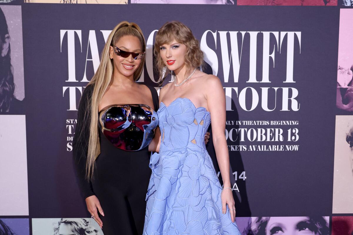 Beyoncé in a black bodysuit with metallic breast plate and sunglasses stands next to Taylor Swift in a blue strapless dress.