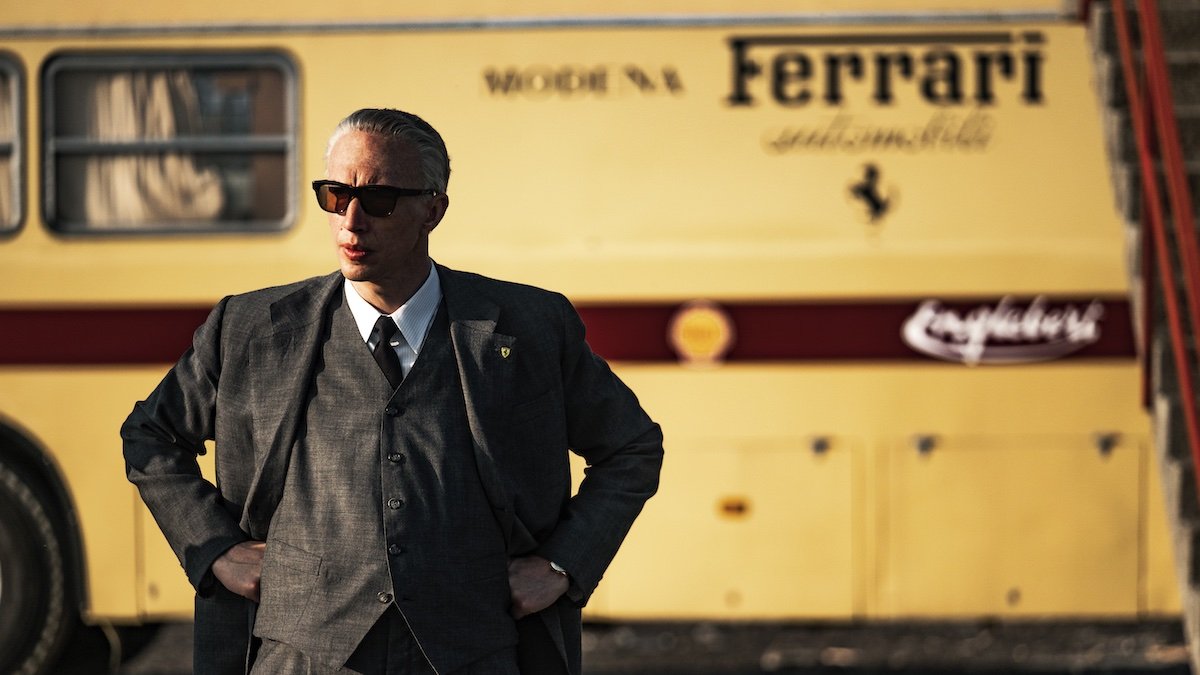 Adam Driver in a three-piece suit as Enzo Ferrari in front of a yellow vehicle with Ferrari on it